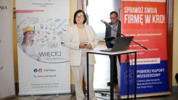 Kobieta przemawia na konferencji stojąc przy podium z laptopem, obok transparentów Powiatowego Urzędu Pracy w Nysie oraz KRD (Krajowy Rejestr Długów).