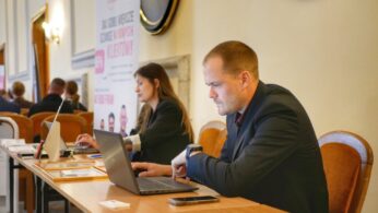 Mężczyzna i kobieta pracują na laptopach przy długim stole podczas wydarzenia biznesowego.