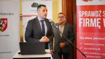 Mężczyzna w garniturze przemawia do mikrofonu, obok stoi drugi mężczyzna. W tle widać baner KRD.