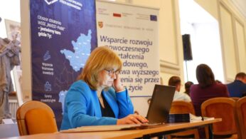 Kobieta w niebieskiej marynarce pracuje na laptopie przy stole, w tle widoczne transparenty dotyczące wsparcia rozwoju i inwestycji.