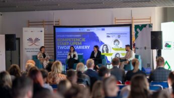 Zdalny widok na scenę, na której panel czterech osób (trzech kobiet i jednego mężczyzny) siedzi na niebieskich kanapach, rozmawiając podczas wydarzenia. Za nimi znajduje się duży ekran z napisem 