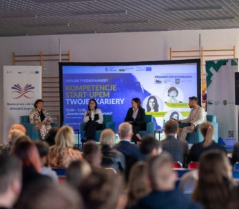 Zdalny widok na scenę, na której panel czterech osób (trzech kobiet i jednego mężczyzny) siedzi na niebieskich kanapach, rozmawiając podczas wydarzenia. Za nimi znajduje się duży ekran z napisem "OPOLSKI TYDZIEŃ KARIERY", "KOMPETENCJE START-UPEM TWOJEJ KARIERY" oraz logotypami organizatorów. Na pierwszym planie widać tyły głów publiczności siedzącej w rzędach, skupionej na scenie.