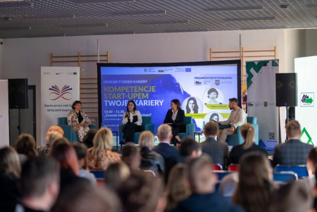 Zdalny widok na scenę, na której panel czterech osób (trzech kobiet i jednego mężczyzny) siedzi na niebieskich kanapach, rozmawiając podczas wydarzenia. Za nimi znajduje się duży ekran z napisem "OPOLSKI TYDZIEŃ KARIERY", "KOMPETENCJE START-UPEM TWOJEJ KARIERY" oraz logotypami organizatorów. Na pierwszym planie widać tyły głów publiczności siedzącej w rzędach, skupionej na scenie.