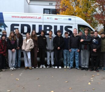 Grupa uczniów i uczennic pozuje przed białym vanem z napisem "EDUBUS" na tle jesiennych drzew.