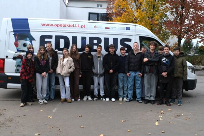 Grupa uczniów i uczennic pozuje przed białym vanem z napisem "EDUBUS" na tle jesiennych drzew.