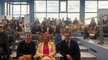 Zdjęcie przedstawia dużą grupę dorosłych osób siedzących w auli z dużymi przeszklonymi oknami. Trzy osoby w pierwszym rzędzie – dwie kobiety i mężczyzna – uśmiechają się do aparatu.