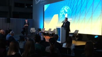 Marszałek województwa opolskiego przemawia na mównicy na scenie podczas konferencji. W tle duży ekran z grafiką i napisem 