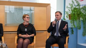 Dwie osoby, kobieta i mężczyzna, siedzą na krzesłach podczas panelu dyskusyjnego. Mężczyzna w garniturze mówi do mikrofonu i gestykuluje, natomiast kobieta w czarnej sukience i okularach słucha. W tle widać herb na szklanej tablicy.