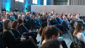 Uczestnicy konferencji siedzą w rzędach na sali, oświetlonej niebieskim światłem z boku.