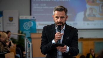 Mężczyzna w ciemnej marynarce w kratę i białej koszuli mówi do mikrofonu, trzymając w drugiej ręce telefon. Tło stanowi rozmyty ekran projekcyjny z napisem 