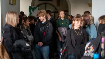 Grupa młodych ludzi stoi w korytarzu lub przejściu o jasnych ścianach i sklepionym suficie. Centralnie widoczny jest chłopak o jasnych, kręconych włosach.