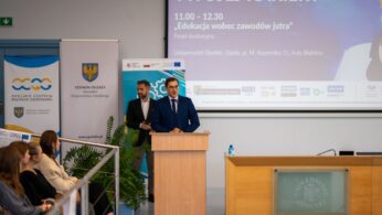 Mężczyzna w granatowym garniturze stoi za drewnianym podium i przemawia, prowadząc panel dyskusyjny 