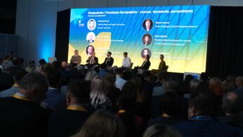 Panel dyskusyjny na scenie podczas konferencji. W tle duży ekran z tytułem 