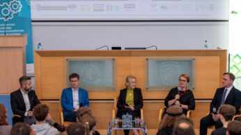Pięcioro prelegentów siedzi na krzesłach na scenie przed publicznością, prowadząc dyskusję panelową. Kobieta w okularach trzyma mikrofon i mówi. Za panelistami widać drewnianą zabudowę i dwie szklane tablice z herbami Uniwersytetu Opolskiego.