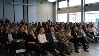 Szerokie ujęcie publiczności podczas konferencji siedzącej w rzędach krzeseł.