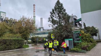 Grupa zwiedzających w żółtych kaskach i kamizelkach idzie na zewnątrz, na terenie zakładu. W tle widoczne są wysokie kominy, zielone budynki przemysłowe i drzewa.