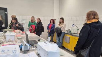 Grupa zwiedzających stoi w nowoczesnym laboratorium o białych ścianach i żółtych szafkach, obserwując sprzęt analityczny na blacie. Na stole widoczne są próbki materiałów, notatki i laptop.