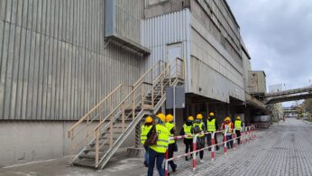 Grupa zwiedzających idzie w rzędzie wzdłuż ściany hali przemysłowej, pod zardzewiałą zewnętrzną klatką schodową. Wszyscy mają na sobie żółte kaski i kamizelki.