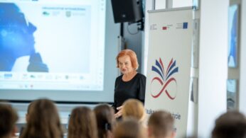 Kobieta z rudymi włosami, w czarnej bluzce, przemawia do widowni zgromadzonej na sali. Z tyłu za nią widać duży ekran projekcyjny z niebieską, graficzną prezentacją oraz baner z napisem w tle.