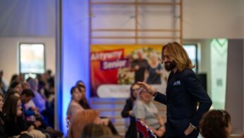 Mężczyzna o długich włosach i brodzie, w ciemnym garniturze, stoi i gestykuluje, przemawiając do publiczności. W tle widać baner promujący 