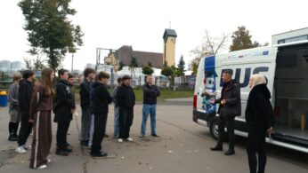 Grupa uczniów słucha mężczyzny stojącego obok białego vana z napisem 