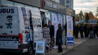 Biały bus EDUBUS z logotypami jest zaparkowany na zewnątrz budynku Toyota Park. Przy busie stoi troje dorosłych ludzi rozmawiających, a w tle widać grupę młodych osób.