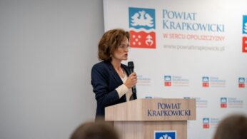 Kobieta w granatowej marynarce przemawia do mikrofonu za mównicą z napisem 