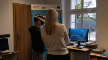Chłopak w goglach VR i kobieta bez gogli, stoją przed monitorem komputera w sali lekcyjnej.