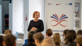 Kobieta z rudymi, krótkimi włosami, ubrana na czarno, stoi i uśmiecha się, przemawiając do zgromadzonej publiczności. Za nią widać baner promocyjny z logo i tekstami dotyczącymi projektów finansowanych, w tym 