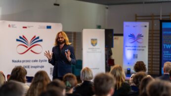 Mężczyzna o długich włosach i brodzie w ciemnym garniturze przemawia do publiczności, energicznie gestykulując obiema rękami. Za nim widoczne są banery z hasłem 