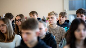 Zbliżenie na twarze młodych uczestników siedzących w rzędach, słuchających prelekcji.