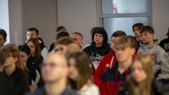 Zbliżenie na fragment publiczności, głównie młodych osób, siedzących na spotkaniu. W centralnej części widać młodego mężczyznę z dredami i opaską na włosach w czarnej bluzie.