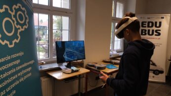 Chłopak używa gogli VR, stojąc przed monitorem komputera, obok okna.