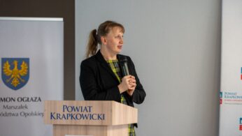 Uśmiechnięta kobieta stoi za mównicą z napisem „Powiat Krapkowicki”.