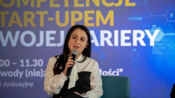 Zbliżenie na kobietę o długich, ciemnych włosach w biało-czarnej bluzce, mówiącą do mikrofonu, siedząc na turkusowym fotelu. W tle widoczny jest baner z hasłem 