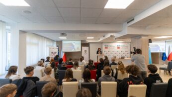sala pełna młodych ludzi, słuchających ciemnowłosej kobiety zza mównicy.