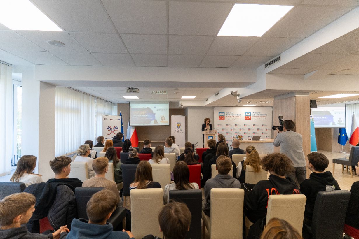 sala pełna młodych ludzi, słuchających ciemnowłosej kobiety zza mównicy.