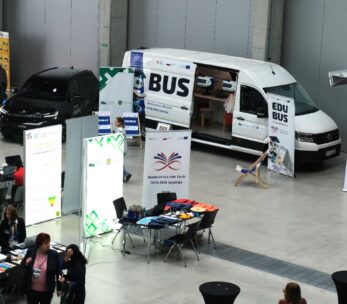 Ogólny widok hali Centrum Wystawienniczo Kongresowego w Opolu z różnymi stoiskami, w tym dużym białym vanem "EDUBUS", dwoma czarnymi samochodami i stoiskami informacyjnymi wystawców Kongresu.