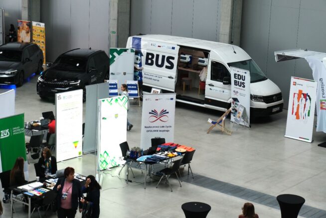 Ogólny widok hali Centrum Wystawienniczo Kongresowego w Opolu z różnymi stoiskami, w tym dużym białym vanem "EDUBUS", dwoma czarnymi samochodami i stoiskami informacyjnymi wystawców Kongresu.