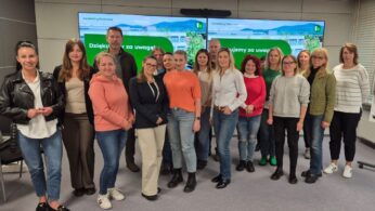 Grupowe zdjęcie kilkunastu uczestników (mężczyzn i kobiet) w sali konferencyjnej. Stoją oni przed ekranami, na których wyświetlone są zielone slajdy z logo Heidelberg Materials i napisem 