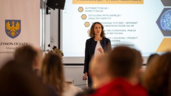 Kobieta o kręconych włosach, ubrana w ciemną marynarkę i białą bluzkę, stoi przed ekranem projekcyjnym, prowadząc prezentację dla widowni.
