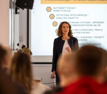 Kobieta o kręconych włosach, ubrana w ciemną marynarkę i białą bluzkę, stoi przed ekranem projekcyjnym, prowadząc prezentację dla widowni.