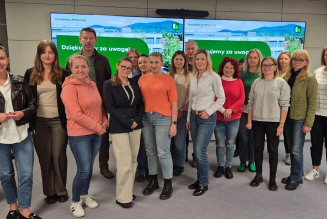 Grupowe zdjęcie kilkunastu uczestników (mężczyzn i kobiet) w sali konferencyjnej. Stoją oni przed ekranami, na których wyświetlone są zielone slajdy z logo Heidelberg Materials i napisem "Dziękujemy za uwagę!".