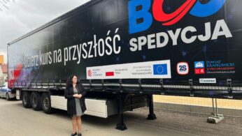 Kobieta w ciemnej marynarce stoi na zewnątrz obok ogromnej naczepy ciężarówki w kolorze czarnym. Na naczepie widnieje duży, czerwono-niebieski napis 