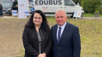 Portretowa poza uśmiechniętej kobiety i starszego mężczyzny w garniturze. Stoją na trawiastym terenie przed białym furgonem EDU-BUS.