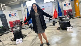 Kobieta w ciemnej marynarce i wzorzystej sukience, z szeroko rozłożonymi rękami, stoi w hali przemysłowej pomiędzy dwoma dużymi dronami. Na dronach widoczne są tabliczki z imionami 