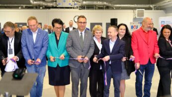Dziewięć osób, oficjeli, ubranych w stroje formalne lub biznesowe, ustawionych w rzędzie, uśmiecha się, trzymając nożyczki i przecinając symboliczną wstęgę w kolorach niebieskim i czerwonym. W tle widać wnętrze dużej, nowoczesnej hali.