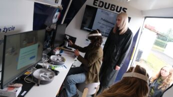 Wnętrze Edubusa. Kilka młodych osób używa sprzętu do wirtualnej rzeczywistości (gogle VR) przy stanowisku komputerowym. Na monitorze widoczny jest tekst podziękowania.