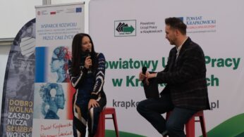 Dwie osoby siedzą na czerwonych stołkach, rozmawiając przy mikrofonach. Kobieta ubrana jest w kombinezon wyścigowy, a mężczyzna w kratkowaną marynarkę. W tle baner z napisem 