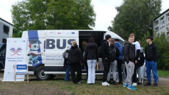 Grupa młodych ludzi stoi wokół otwartego białego busa - EDUBUS, na którego boku widać logo projektu edukacyjnego i hasło w języku polskim. W tle drzewa i budynek.
